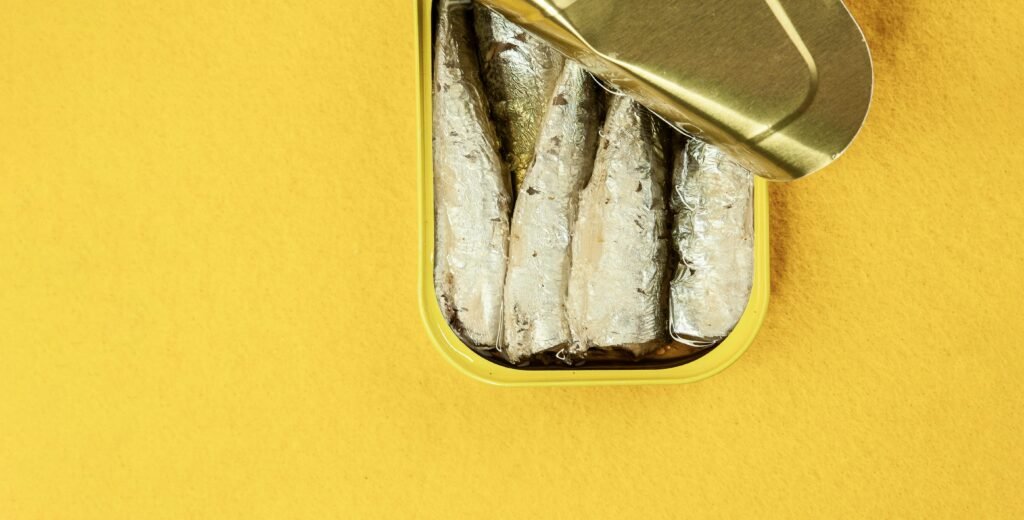 Top view of canned sardines with an open lid on a yellow surface, showcasing food preservation.